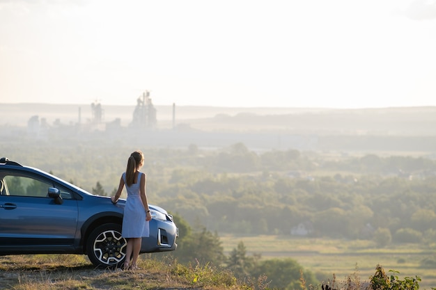 Gelukkig jonge vrouw chauffeur in blauwe jurk genieten van warme zomeravond naast haar auto. Reizen en vakantie concept.