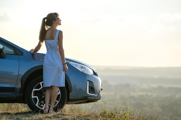 Gelukkig jonge vrouw chauffeur in blauwe jurk leunend op haar auto genieten van warme zomerdag Reizen en vakantie concept