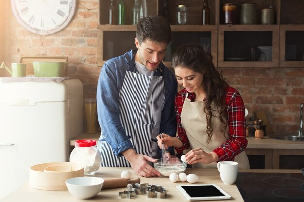 Gelukkig jonge vrouw en man taart bakken in loft keuken. jonge familie koken thuis, met behulp van digitale tablet. mockup voor recept