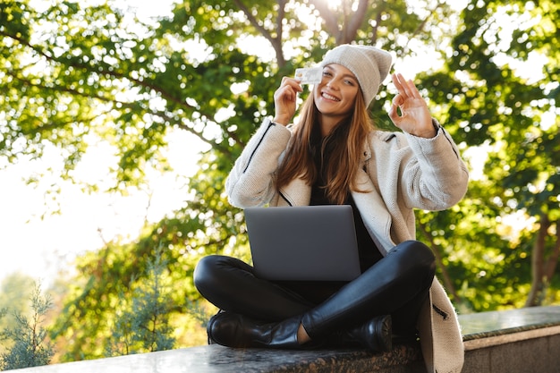 Gelukkig jonge vrouw gekleed in herfst jas en hoed buiten zitten, met behulp van laptop computer, met plastic creditcard