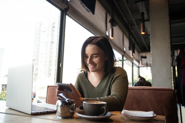 Gelukkig jonge vrouw in café glimlacht op zoek naar smartphone.