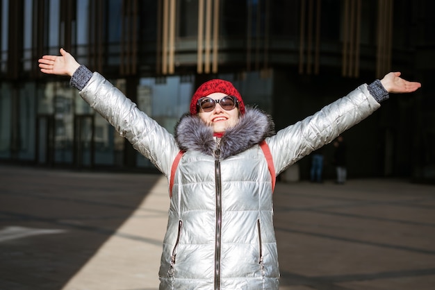 Gelukkig jonge vrouw in zonnebril in een winterjas met een rugzak in de buurt van het gebouw. Reis concept