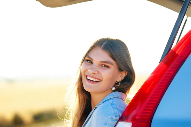 Gelukkig jonge vrouw kijken camera in de buurt van auto bij gouden zonsondergang in veld