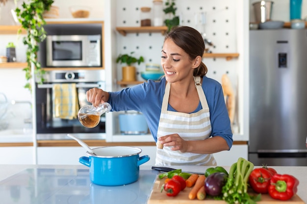 Gelukkig jonge vrouw koken proeverij diner in een pot staande in moderne keuken thuis huisvrouw bereiden van gezonde voeding glimlachend huishouden en voeding dieet recepten concept