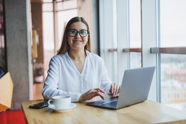 Gelukkig jonge vrouw manager in casual kleding en bril glimlacht en kijkt in een laptop kijkend door verschillende informatie, ze drinkt koffie in een gezellig café met grote ramen en uitzicht op de stad