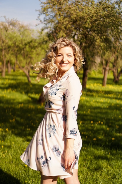 Gelukkig jonge vrouw met blond haar, gekleed in een jurk, buiten poseren in een tuin met kersenbomen in de zon, glimlachend. Krullen die fladderen in de wind