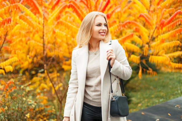 Gelukkig jonge vrouw met een handtas in een trendy jas poseren in de buurt van herfst geel gebladerte