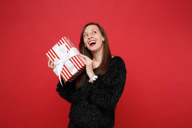 Gelukkig jonge vrouw opzoeken met rood gestreepte huidige doos met cadeau lint geïsoleerd op rode achtergrond. St. Valentijnsdag, Internationale Vrouwendag, verjaardag, vakantieconcept. Bespotten kopie ruimte.