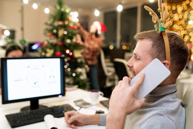 Gelukkig jonge zakenman met xmas hoofdband notities maken tijdens het gesprek op de mobiele telefoon achter computer door werkplek