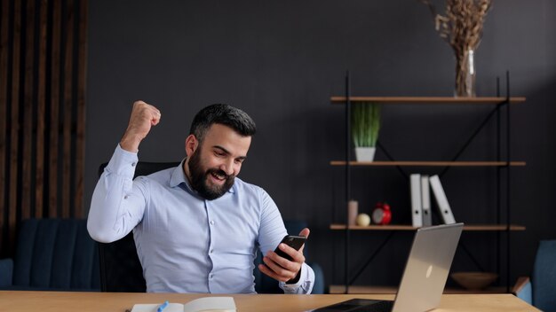 Gelukkig jonge zakenman verrast door het goede nieuws en ja gebaar maken tijdens het kijken naar smartphone zittend op kantoor.