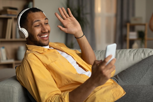 Gelukkig jonge zwarte man in headset communiceren op smartphone zwaaien naar webcam zittend op de bank thuis