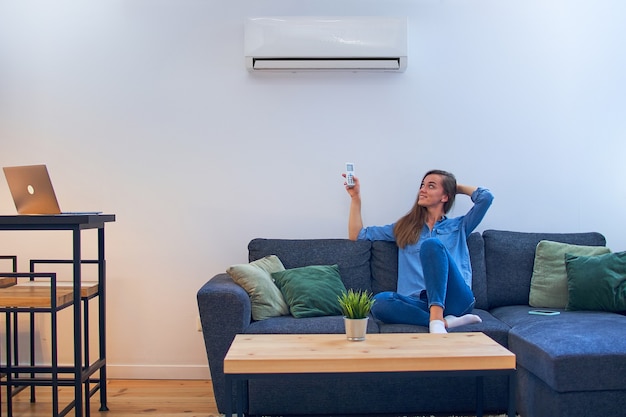 Gelukkig jongedame zittend op de Bank onder airconditioner en comfort temperatuur met afstandsbediening bij modern huis aanpassen