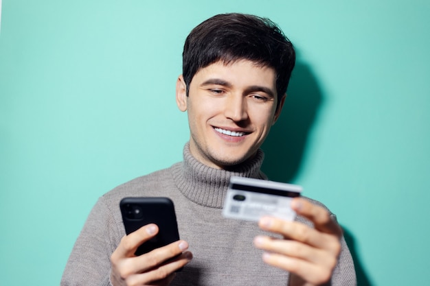 Gelukkig jongeman met smartphone en creditcard in de hand op muur van aqua menthe kleur.