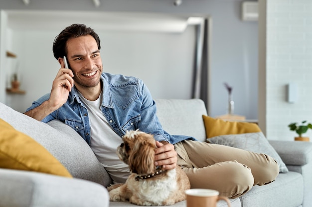 Gelukkig jongeman ontspannen op de bank met zijn hond tijdens het bellen via mobiele telefoon