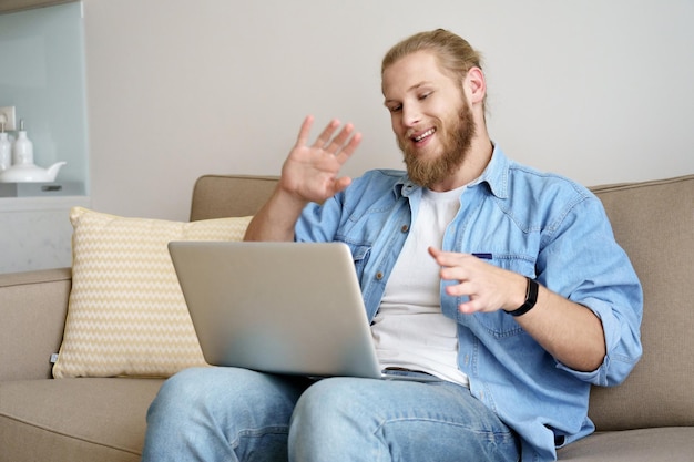 Gelukkig jongeman zwaaiende hand videobellen op laptop zittend op de bank werken studeren vanuit huis extern kantoor glimlachende mannelijke afstandswerker student doet online conferentie webcam chat op computer