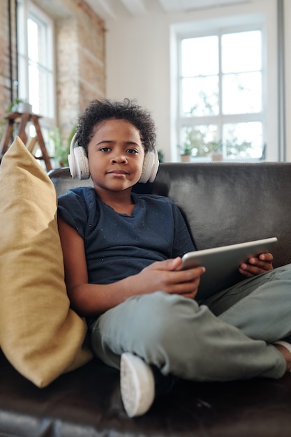 Gelukkig jongetje met koptelefoon zittend op een zwarte leren bank voor de camera en kijken naar online video in tablet in thuisomgeving