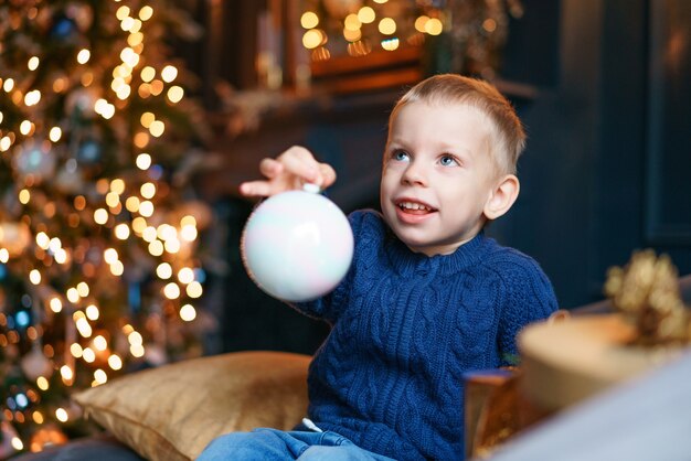 Gelukkig jongetje zittend op de bank met kerstboomversiering in een gezellige woonkamer in blauw gebreide ...