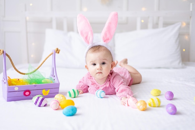 Gelukkig Kaukasisch babymeisje met een rand van konijnenoren op haar hoofd ligt op het bed thuis in de slaapkamer met kleurrijke paaseieren paasbaby