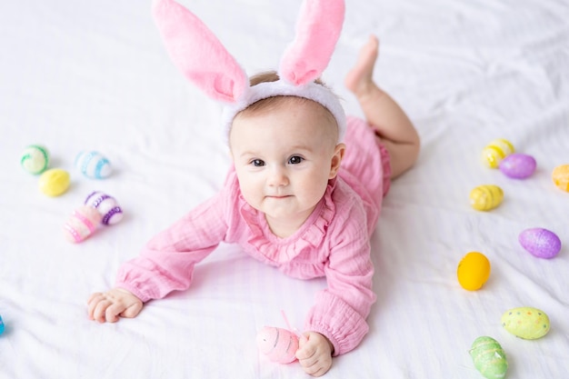 Gelukkig Kaukasisch babymeisje met een rand van konijnenoren op haar hoofd ligt op het bed thuis in de slaapkamer met kleurrijke paaseieren paasbaby