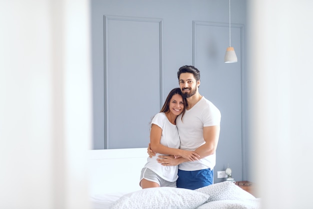 Gelukkig Kaukasisch paar in pyjama's glimlachen, knuffelen in bed en camera kijken. Ochtend tijd. Slaapkamer interieur.