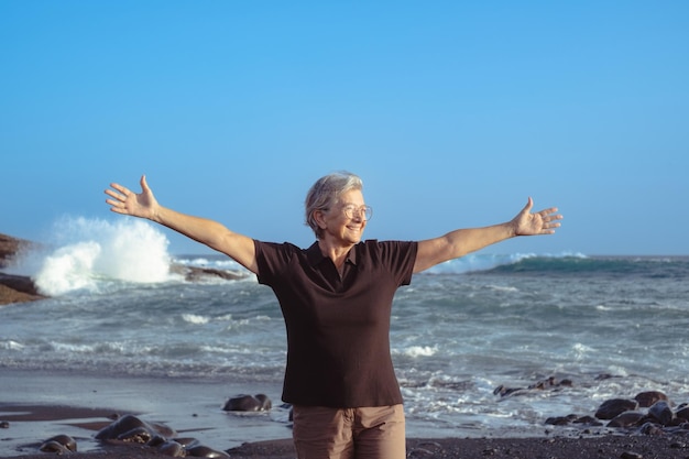 Gelukkig Kaukasische senior vrouw met uitgestrekte armen staande op het strand bij zonsondergang terwijl kijken naar de horizon golven en ruwe zee op de achtergrond