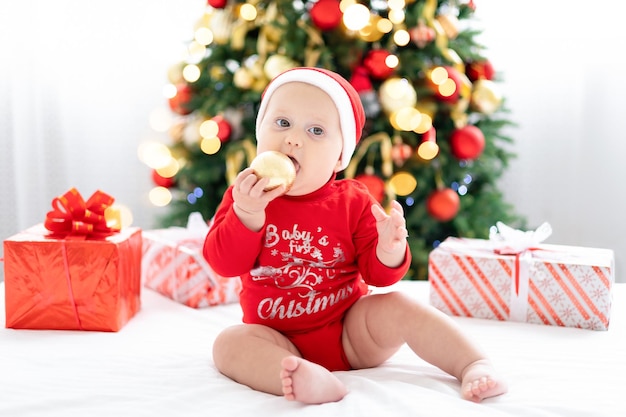 Gelukkig kind baby in rood santa kostuum nieuwjaar thuis vieren met kerstboom en geschenken, concept van vakantie, vrolijk kerstfeest en nieuwjaar