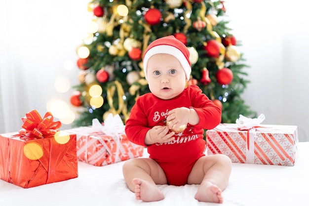 Gelukkig kind baby in rood santa kostuum nieuwjaar thuis vieren met kerstboom en geschenken, concept van vakantie, vrolijk kerstfeest en nieuwjaar