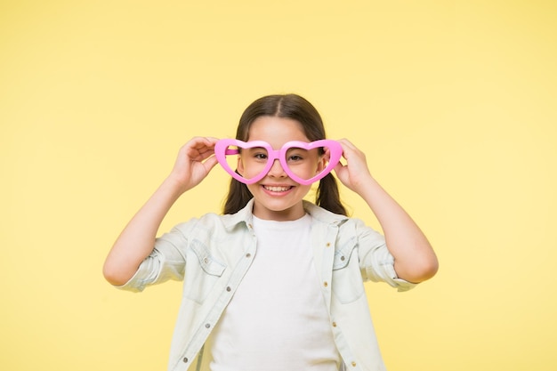 Gelukkig kind draagt een hartvormige bril op gele achtergrond Kleine meisjesglimlach in modeaccessoire Zomermode-look Funky stijl schoonheid Jong en zelfverzekerd Valentijnsdag Ik hou van je