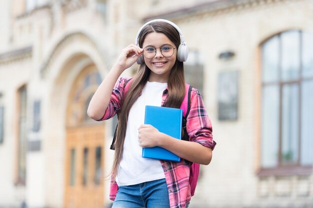 Gelukkig kind in koptelefoon houdt schoolboek vast dat brillen vasthoudt buiten audiocursussen