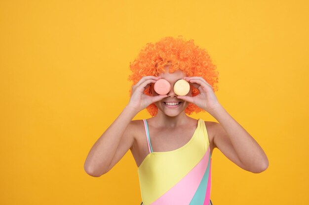 Gelukkig kind in zwembroek met oranje krullend pruikhaar met franse macaron op gele achtergrond, zoetekauw.