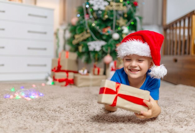 Gelukkig kind jongen op kerstmis op gezellig tapijt in zijn kamer in pyjama en rode kerstmuts open geschenken