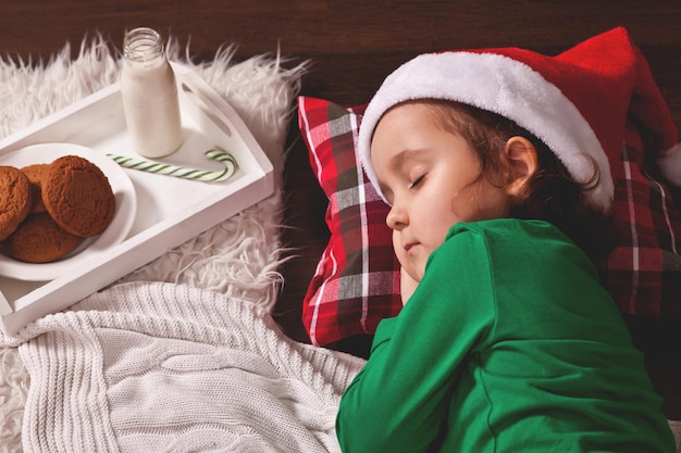 Gelukkig kind meisje in kerstmuts wachten op de kerstman en slapen in de buurt van dennenboom op kerstavond