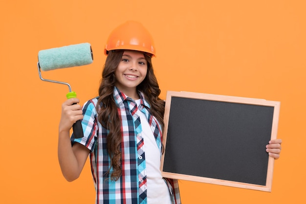 Gelukkig kind met krullend haar in helm houdt verfroller vast en schoolbord kopieerruimte school