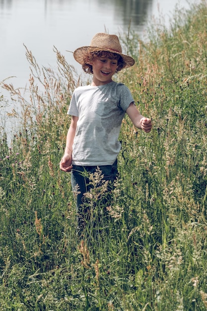 Gelukkig kind met krullend haar met een strohoed die tussen het hoge groene gras in de buurt van het kanaal staat en geniet van een zonnige zomerdag terwijl hij wegkijkt