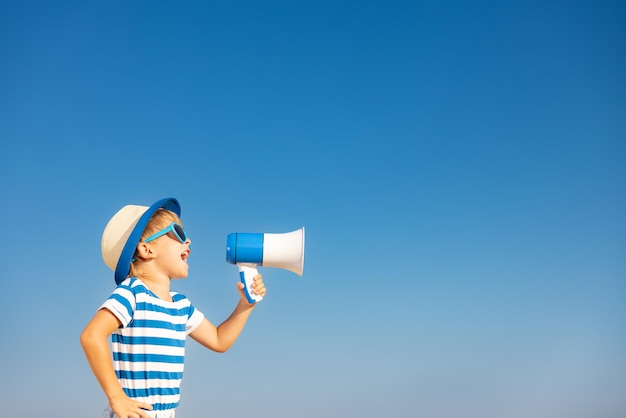 Gelukkig kind schreeuwen door luidspreker buiten tegen blauwe hemelachtergrond. Portret van lachende jongen op zomervakantie. Vrijheid en reisconcept