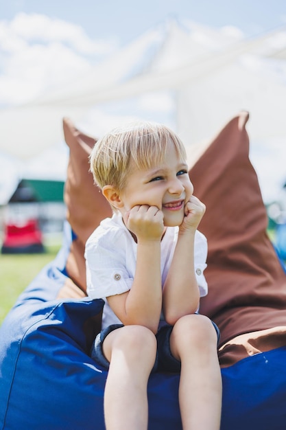 Gelukkig kind zit in een zitzakstoel in het zomerpark Een kleine jongen in een wit T-shirt heeft plezier buitenshuis Jeugd en geluk