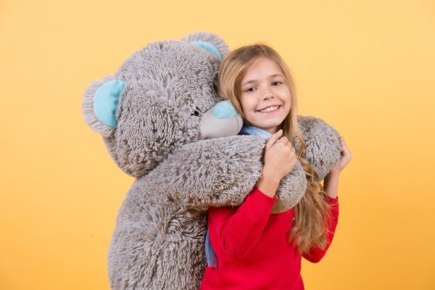 Gelukkig kindertijdconcept. Kid met dierenpop, cadeau en cadeau. Kindglimlach met grijs zacht stuk speelgoed. Meisje knuffel grote teddybeer op oranje achtergrond. Vakantie, verjaardag, jubileumviering.