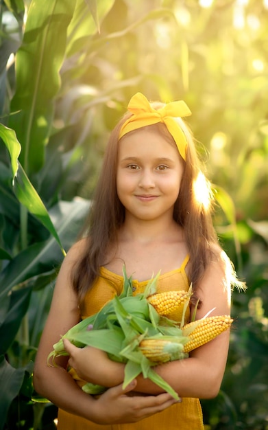 gelukkig kindmeisje staat in het struikgewas van maïs en houdt een gele maïskolf in haar handen