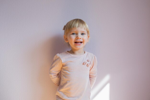 Gelukkig klein blond meisje staat leunend tegen de muur in een zonnige kamer thuis