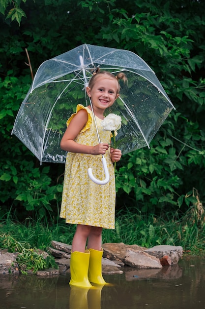 Gelukkig klein emotioneel meisje met transparante paraplu in gele jurk en regenlaarzen in de zomer van het park