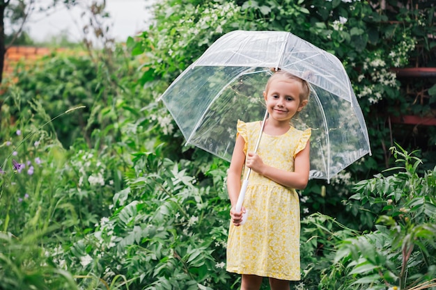Gelukkig klein emotioneel meisje met transparante paraplu in gele jurk en regenlaarzen in de zomer van het park