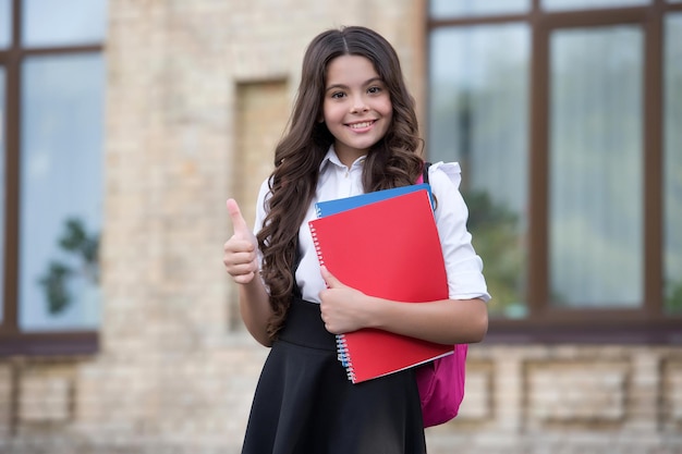 Gelukkig klein kind in back to school-stijl geeft duimen op handgebaar buitenshuis, goedgekeurd.