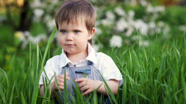 Gelukkig klein kind in modieuze blauwe jumpsuit met mooie blauwe ogen. Grappige speelt in hoog groen gras in een bloeiend park vol groen tegen de achtergrond van een appelboom.