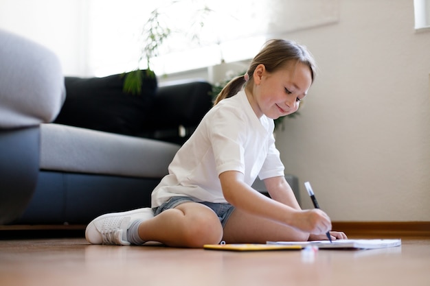 Gelukkig klein kind zit comfortabel op de houten vloer, tekenen op papier met kleurpotloden