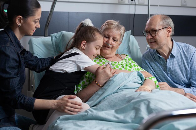 Gelukkig klein meisje begroet zieke grootmoeder met knuffel op bezoek aan ziekenhuiskamer. Oude dame die op een medisch bed ligt en gelukkig knuffels geeft aan kleindochter in de medische kliniek van geriatrische patiënten.