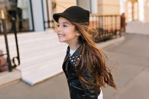 Gelukkig klein meisje dat door de straat rent met donker haar dat zwaait en wegkijkt. portret van een speelse brunette girlie met een zwarte hoed en een stijlvol jasje die tijdens vakanties op de achtergrond van de stad springt