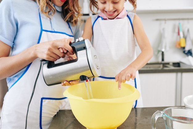 Gelukkig klein meisje en haar moeder die pannenkoekdeeg in grote plastic kom mengen met elektronische mixer