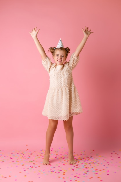 Gelukkig klein meisje in een verjaardag-pet blaast haar handpalmen veelkleurige confetti op een roze achtergrond in de studio