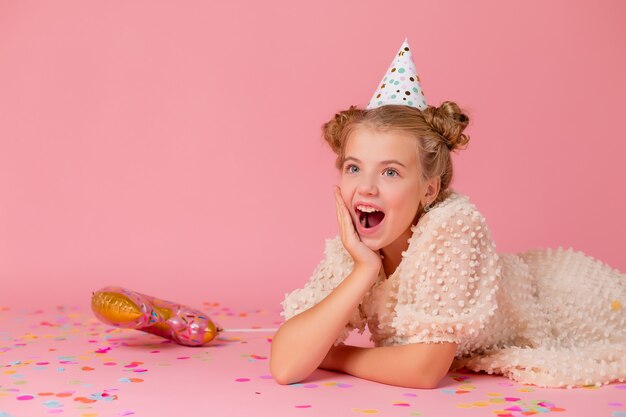 Gelukkig klein meisje in een verjaardag-pet ligt op een roze achtergrond met confetti, houdt haar handen op het gezicht.