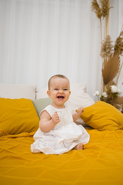 Gelukkig klein meisje in een witte jurk zit op een gele deken op het bed in de slaapkamer